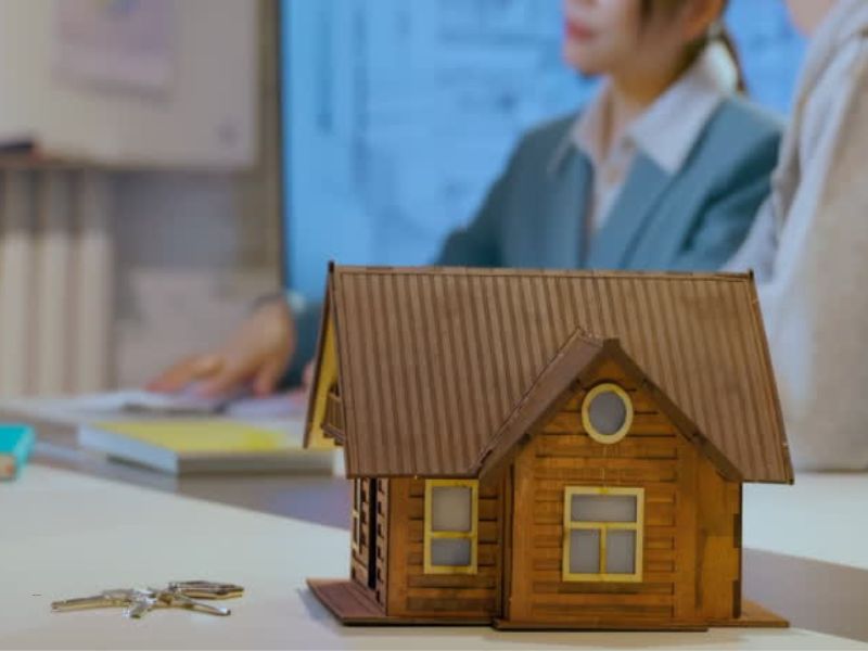 prikaz kuće i ključa u prvom planu, u pozadini tri osobe prveravaju kupoprodajni ugovor