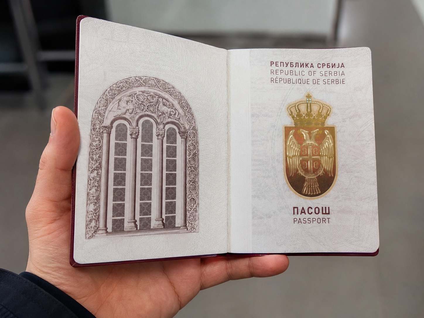 Ruka drži otvoren srpski pasoš sa grbom Republike Srbije i unutrašnjom stranicom dokumenta.